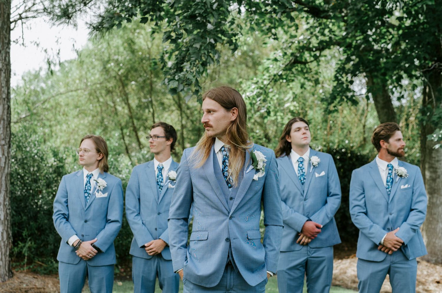 Wedding party members in light blue suits laugh together while standing outdoors in a natural setting.
