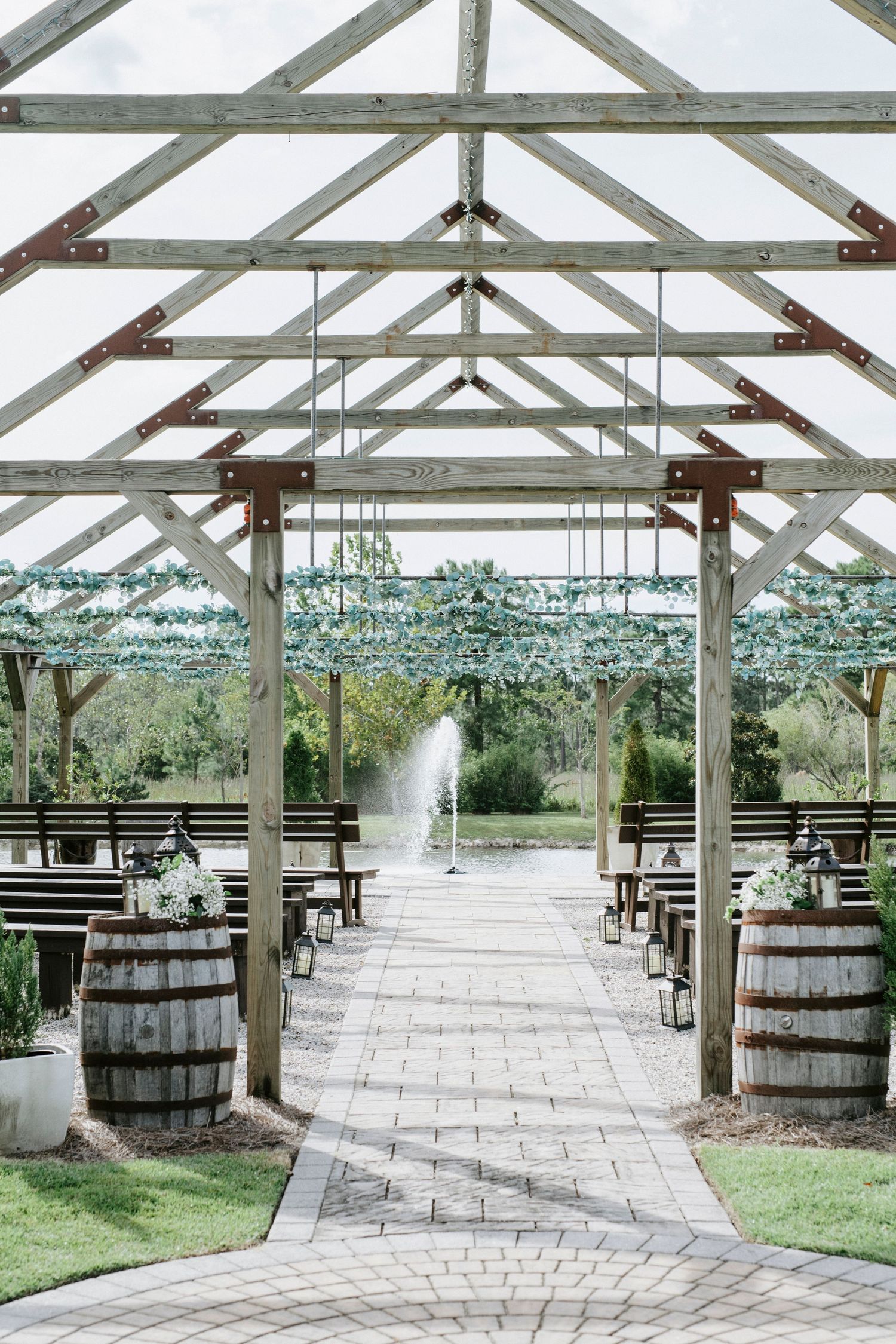 Rustic outdoor wedding ceremony setup with wooden beams, wine barrels and a stone pathway leading to a fountain.