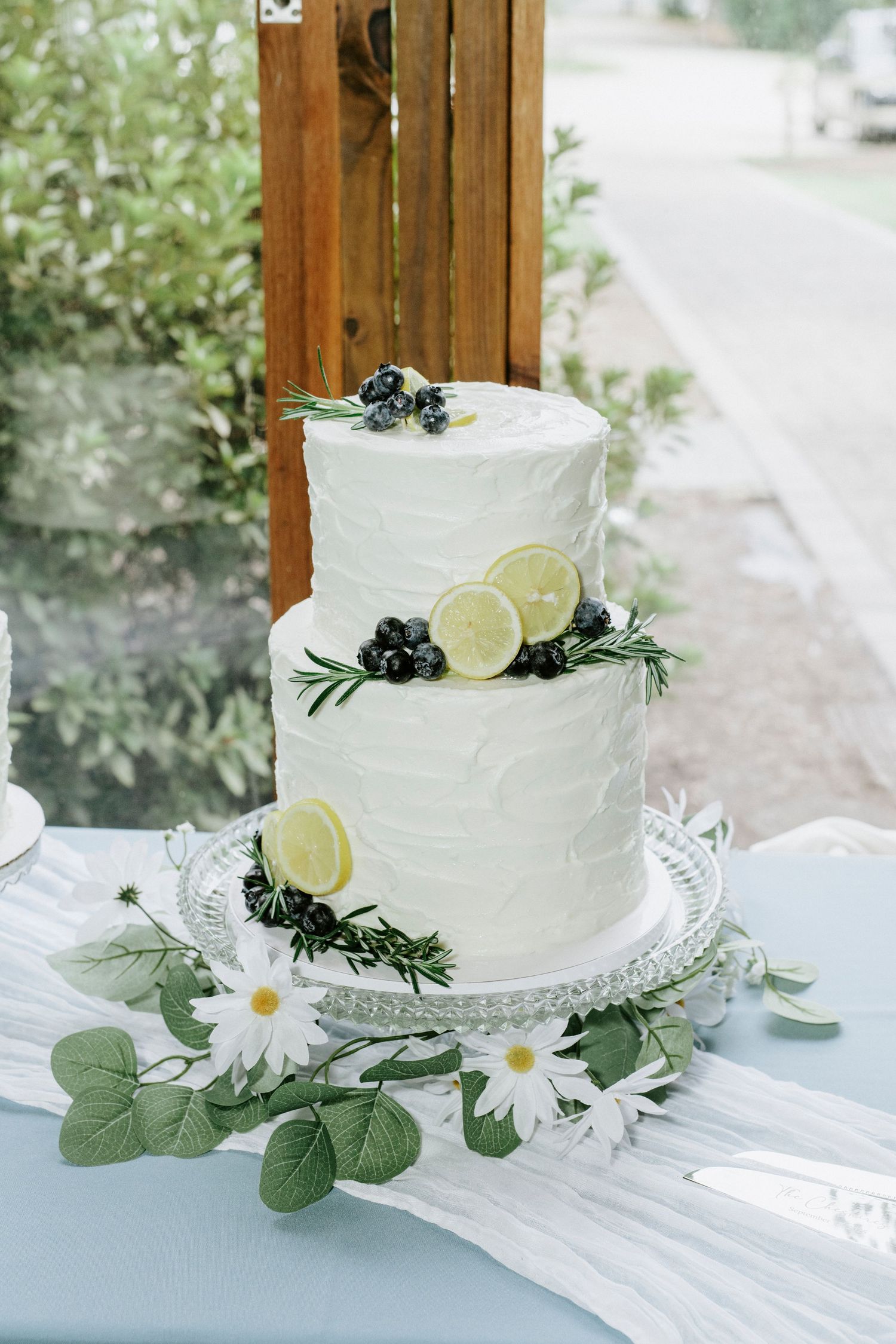 White buttercream wedding cake decorated with fresh daisies, lemon slices, and eucalyptus leaves on a lace doily.