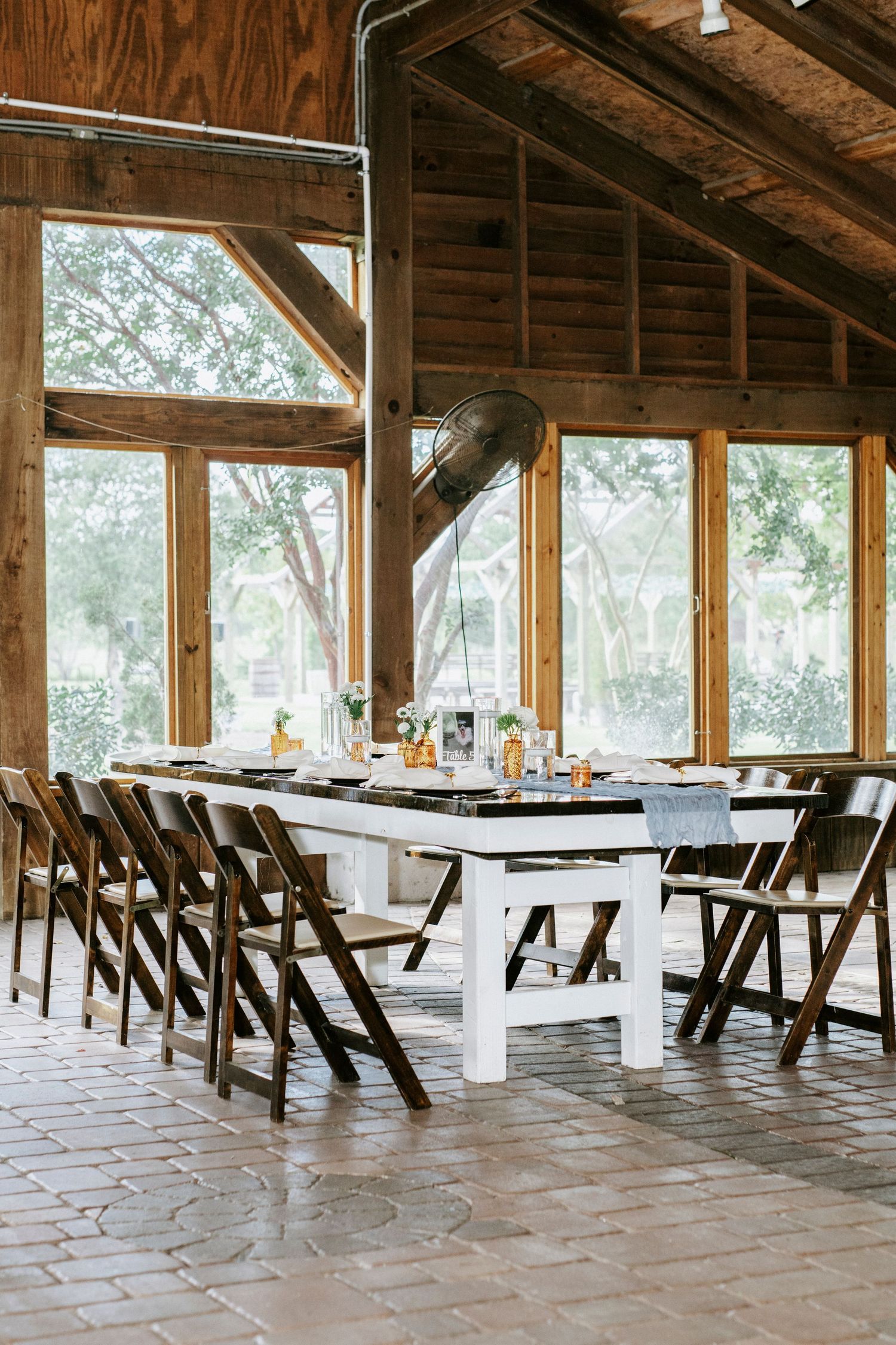 Rustic wooden event venue with long white tables and dark wooden chairs set up under exposed beam ceiling and windows.