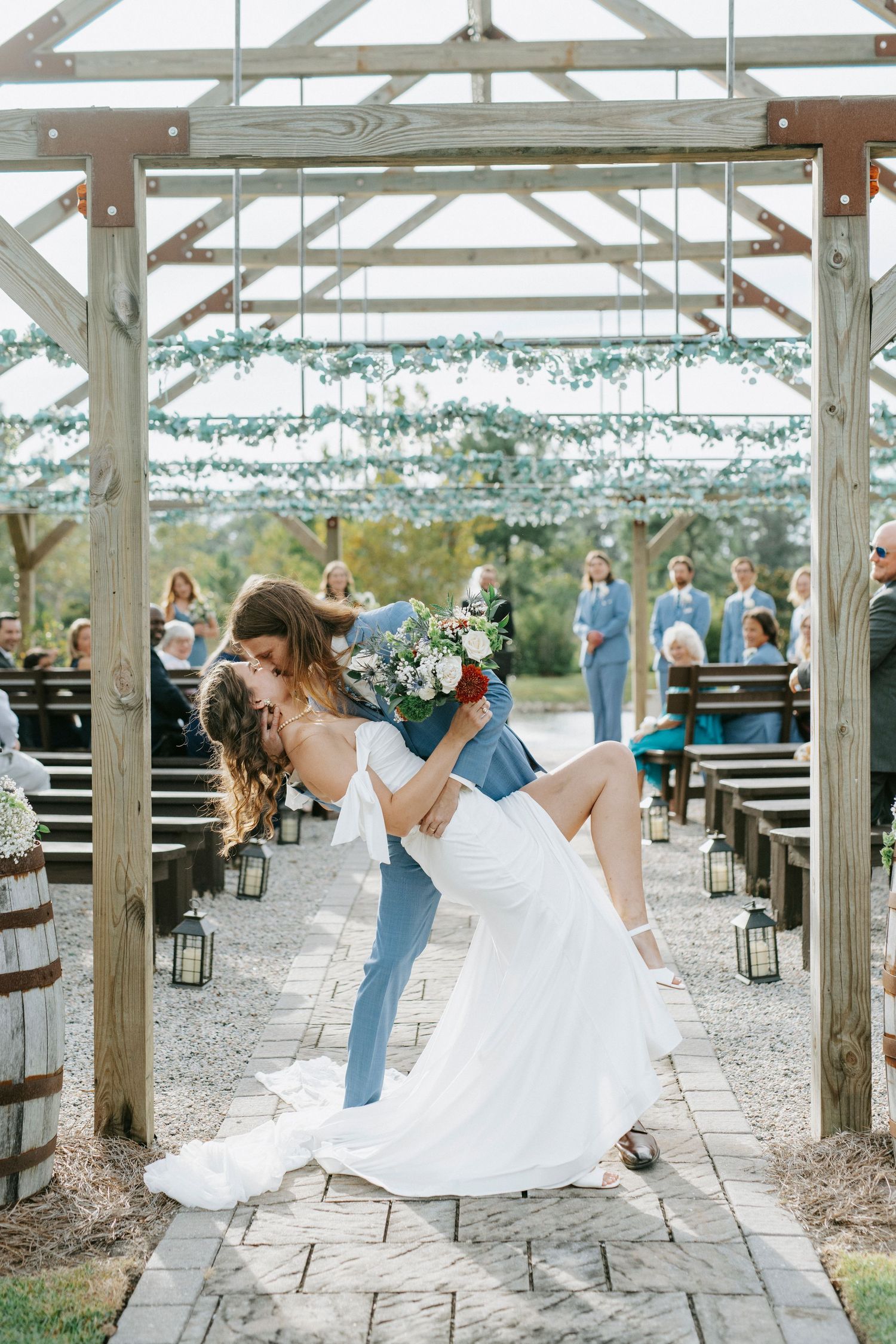 A romantic wedding couple dances and dips together under a white wooden pergola decorated with string lights.