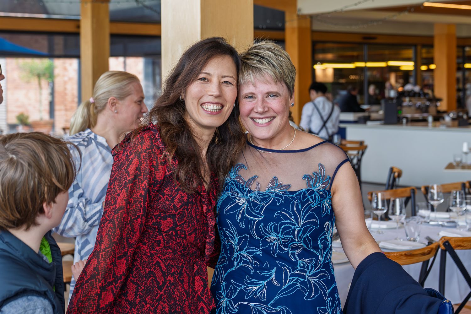 Two smiling women embrace at a waterfront restaurant dining event, wearing colorful patterned outfits.