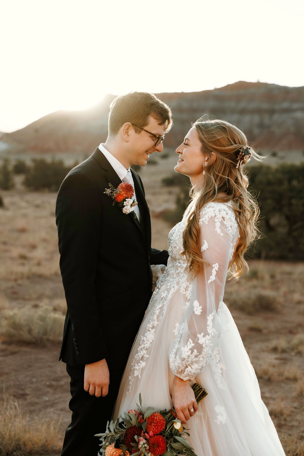 Laughing bride in lace wedding gown and groom in black tuxedo share romantic moment against New Mexico desert sunset backdrop.