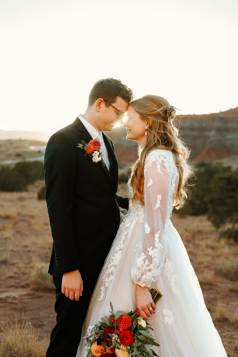 Bride and groom touching foreheads at golden hour in New Mexico desert landscape, bride holding vibrant red bouquet in lace gown.