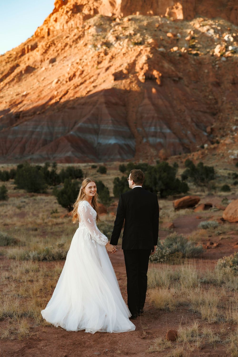 New Mexico Bride in lace gown holds groom's hand walking toward red rock canyon formations during golden hour desert shoot.