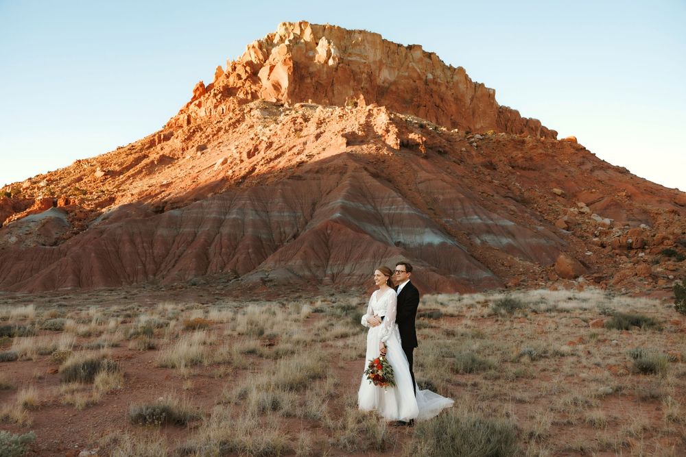New Mexico Bride and groom pose together before a dramatic red sandstone butte in a southwestern desert landscape at dusk.