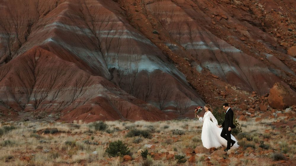 Abiquiu Newlyweds walk hand in hand through a striking red and New Mexico gray striped desert canyon landscape at sunset.