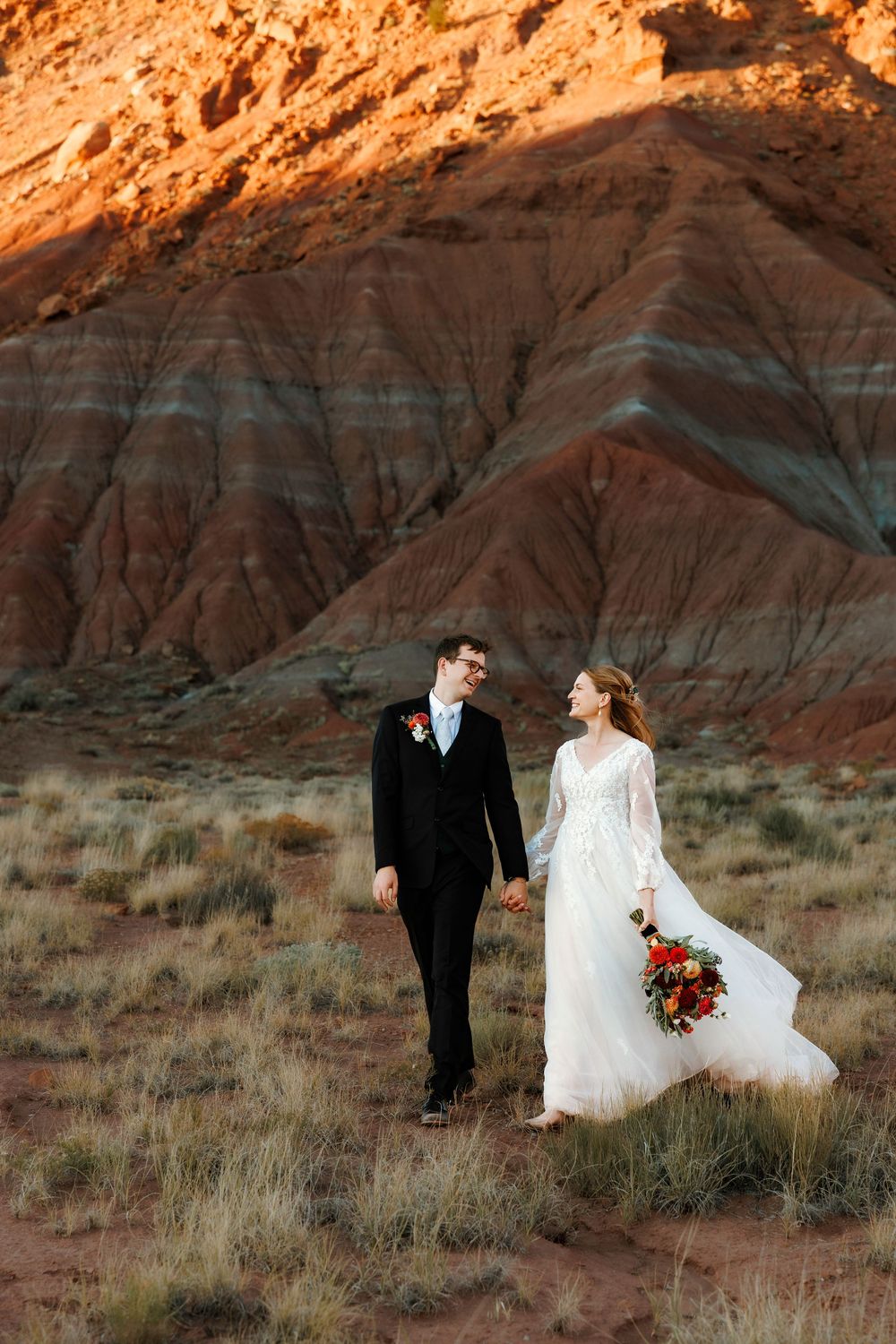 Bride and groom walk hand in hand laughing before layered Abiquiu red rock formations, bride holding autumn bridal bouquet.