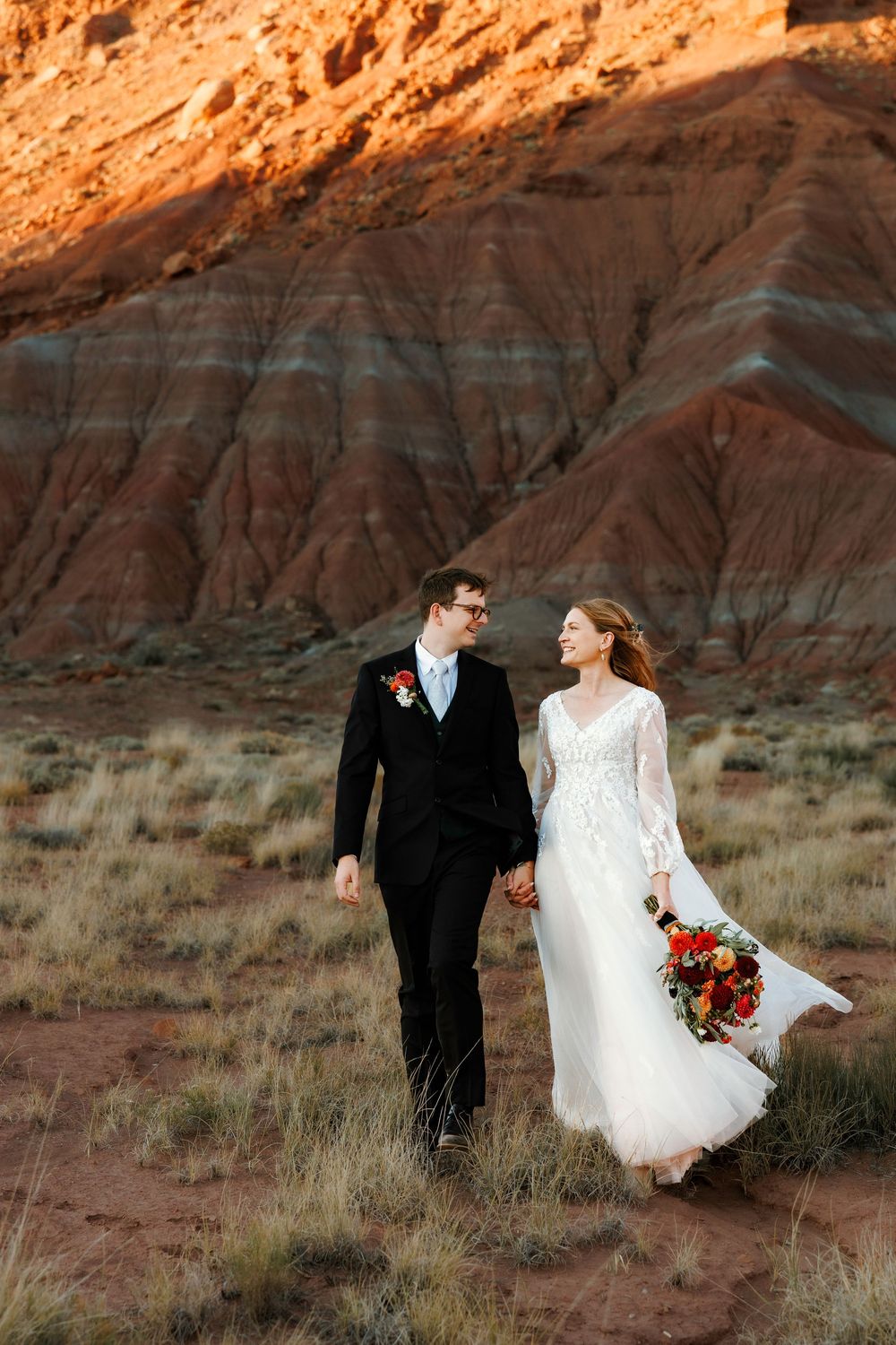 Newlyweds stroll hand in hand through desert terrain with vibrant Abiquiu red striped rock formations glowing at sunset.
