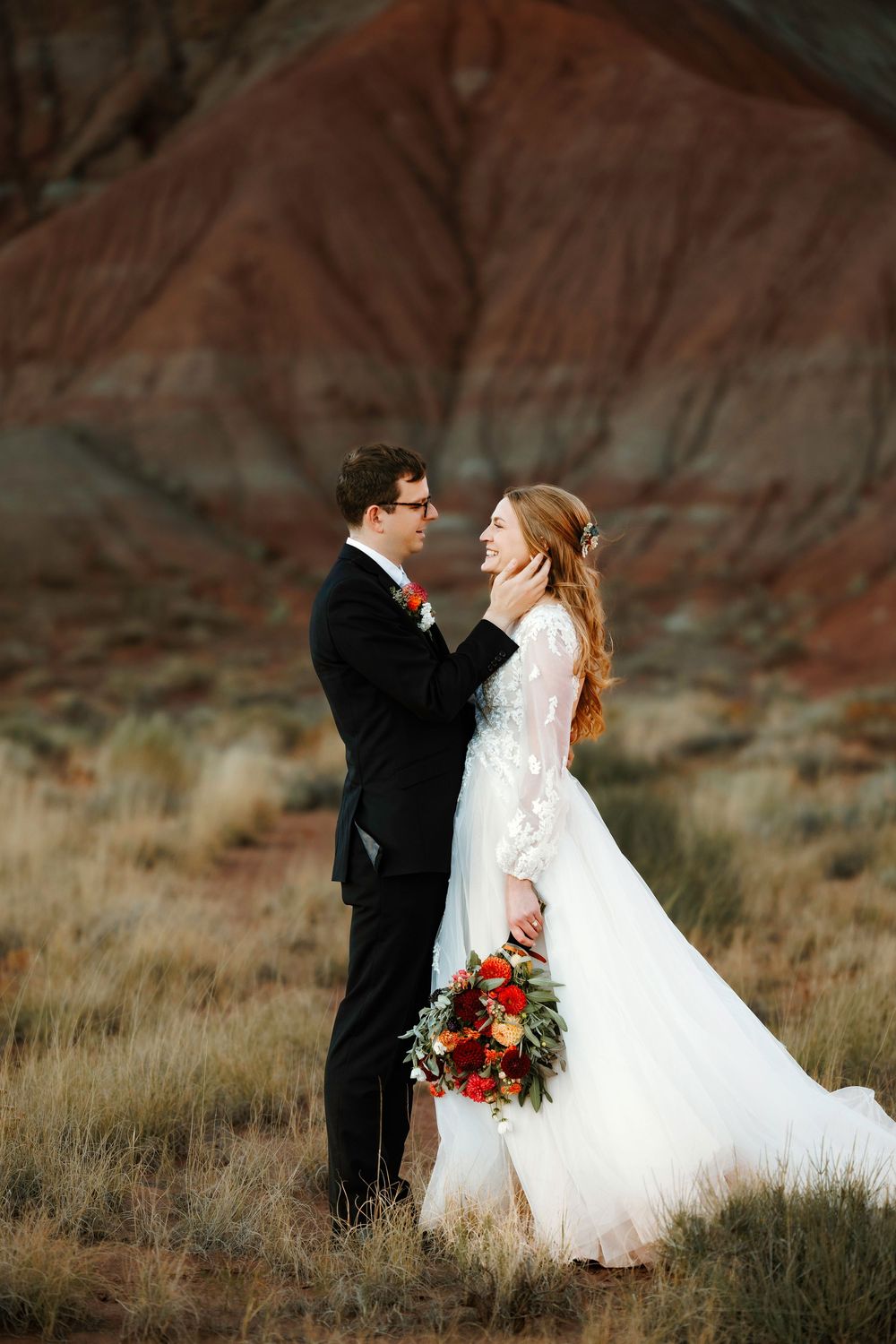 Groom tenderly touches bride's face as she laughs joyfully, standing before Abiquiu red desert hills with bridal bouquet.