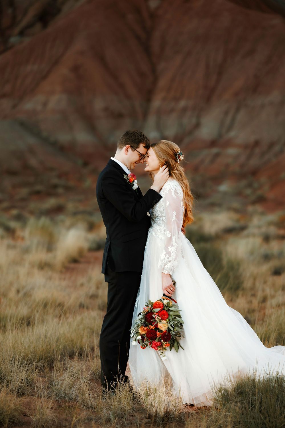 Bride and groom share an intimate forehead touch in golden desert light with red rock formations behind them.