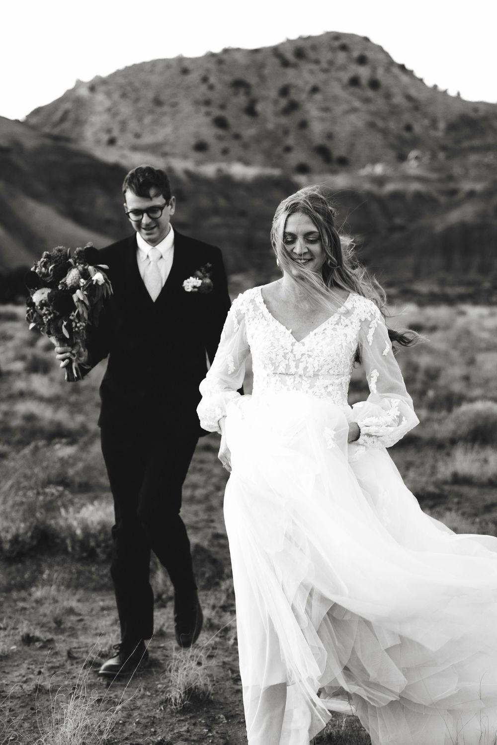Black and white photo of bride and groom walking outdoors with mountain backdrop, bride's dress flowing in wind.