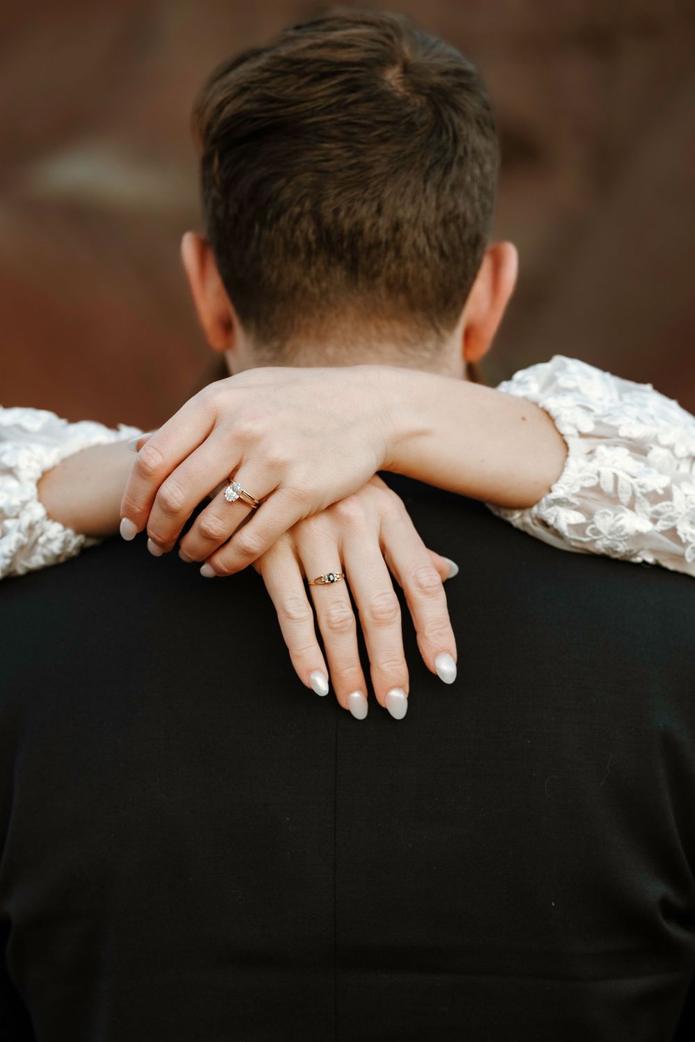 Close-up of bride's hands with wedding ring resting on groom's shoulders from behind.