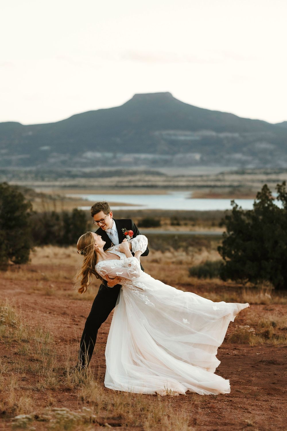 Abiquiu Groom dipping bride in flowing white dress with mountain lake landscape and Pedernal backdrop