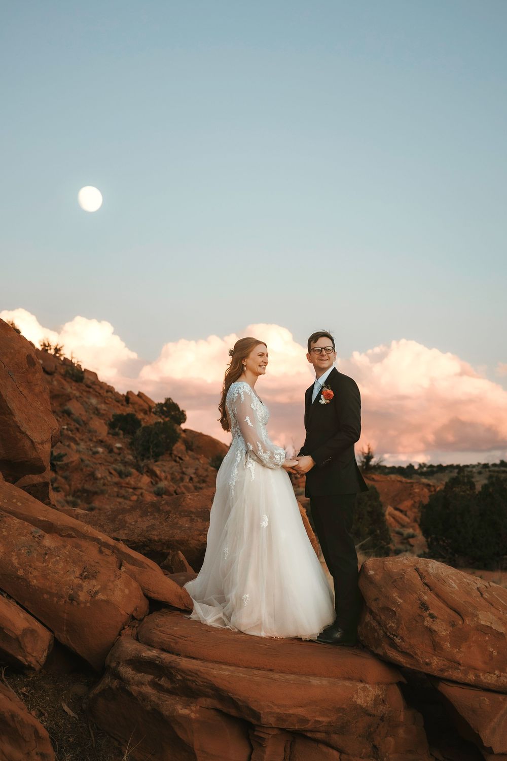 Abiquiu bride and groom holding hands on New Mexico red rocks at dusk with full moon and pink clouds above.