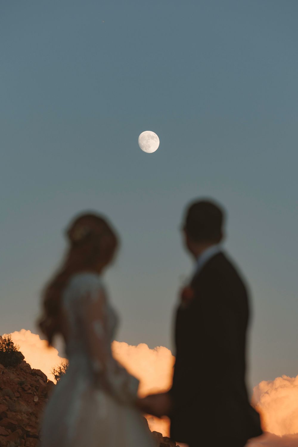 Romantic blurred silhouette of bride and groom holding hands with full moon in twilight sky.