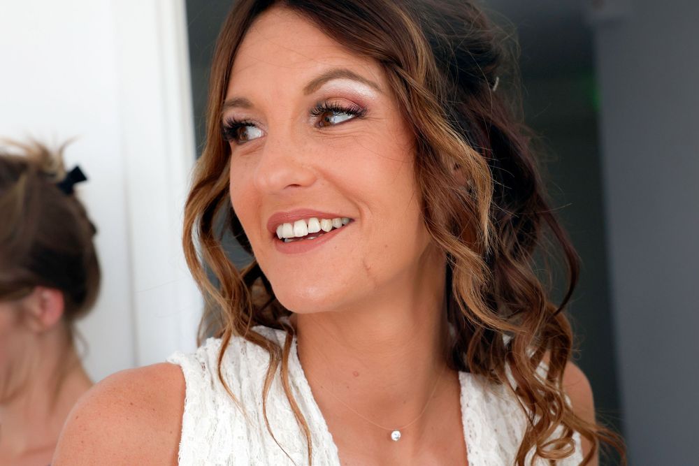 A smiling person with wavy brown hair and natural makeup wearing a white top and delicate necklace.