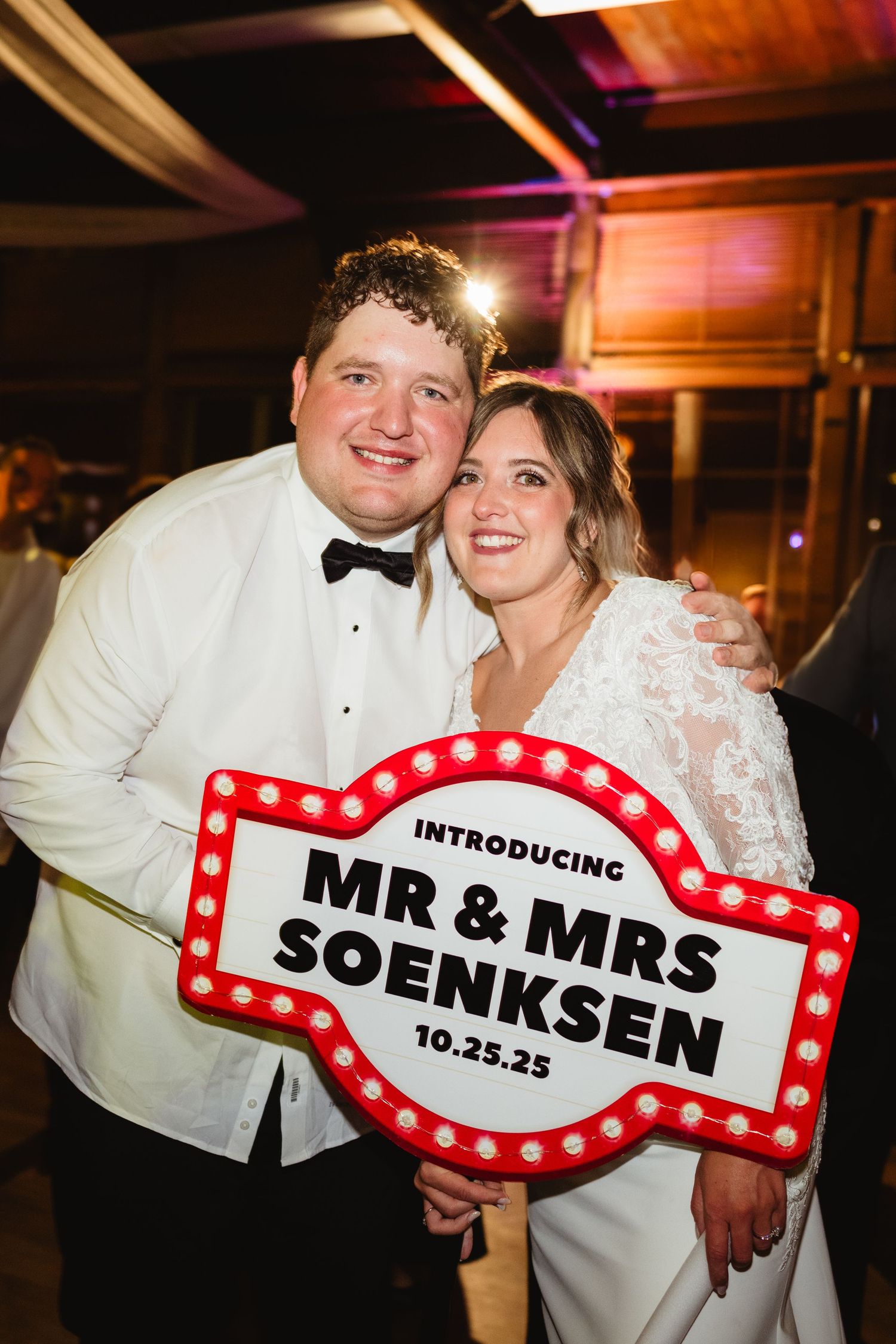 Newlywed couple poses with red and white marquee-style signs displaying their new married name at wedding reception.