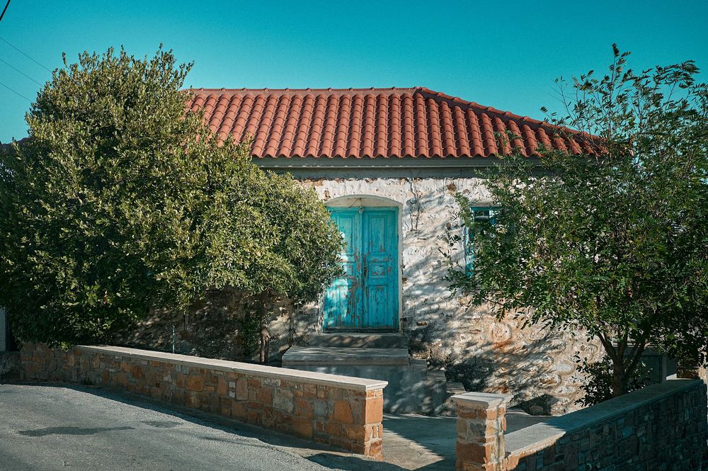 Mediterranean stone house with terracotta roof tiles and turquoise door nestled among green shrubs.