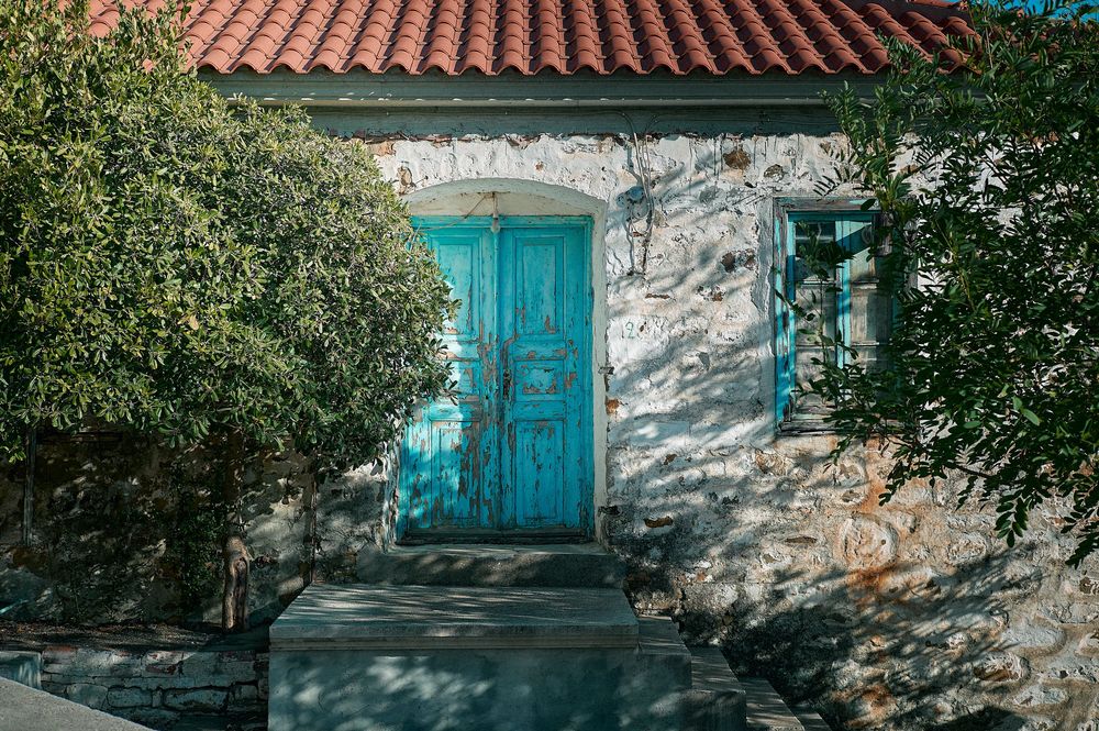 A weathered stone house with bright turquoise door and window frames surrounded by green foliage.