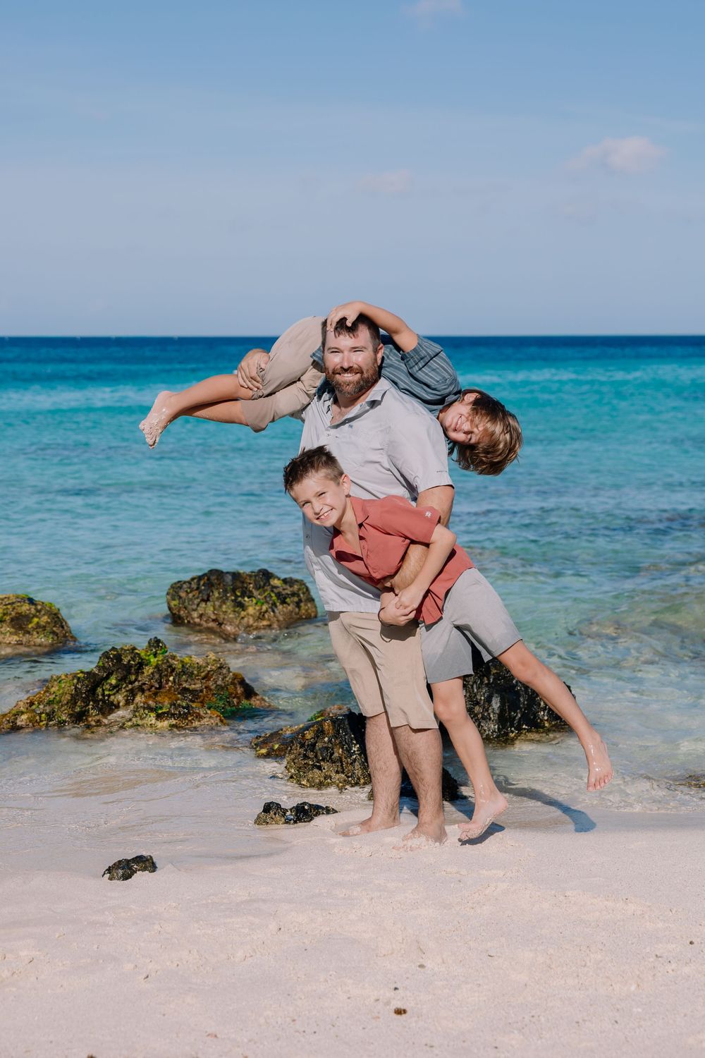 Family plays together on a tropical beach with turquoise water and rocky outcrops in the background.
