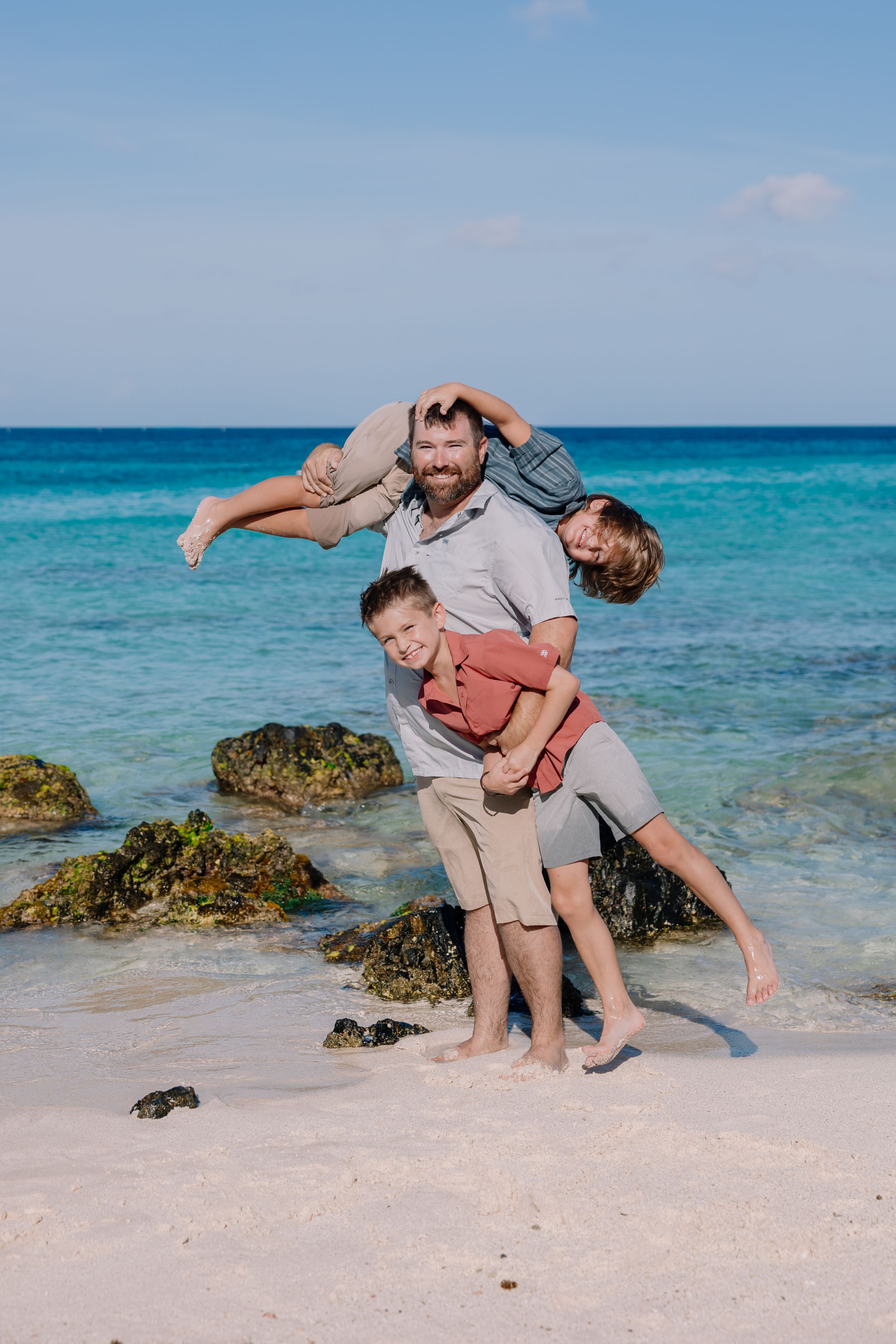 Family plays together on a tropical beach with turquoise water and rocky outcrops in the background.