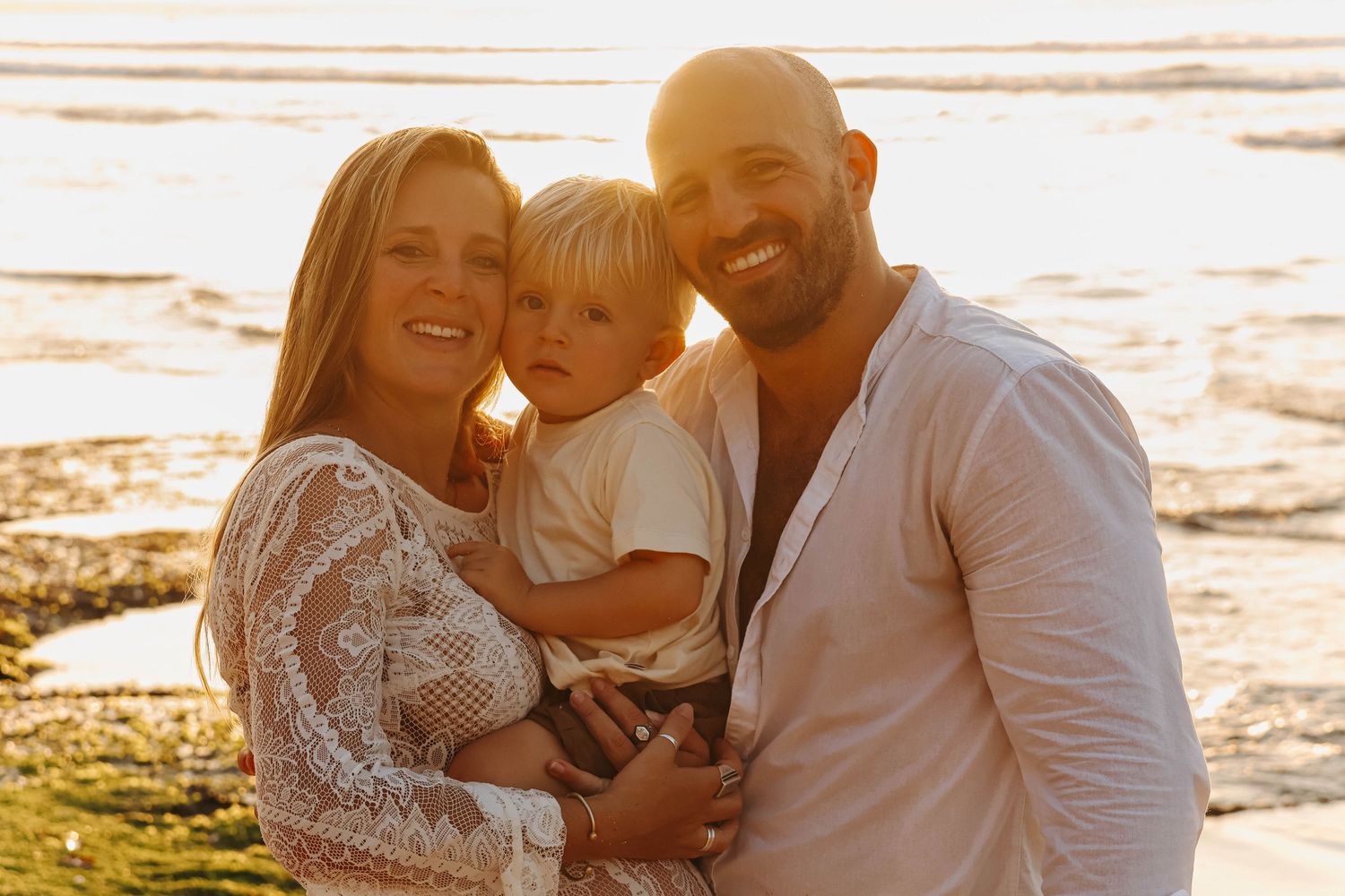 Happy family with toddler posing together on beach at warm golden sunset.