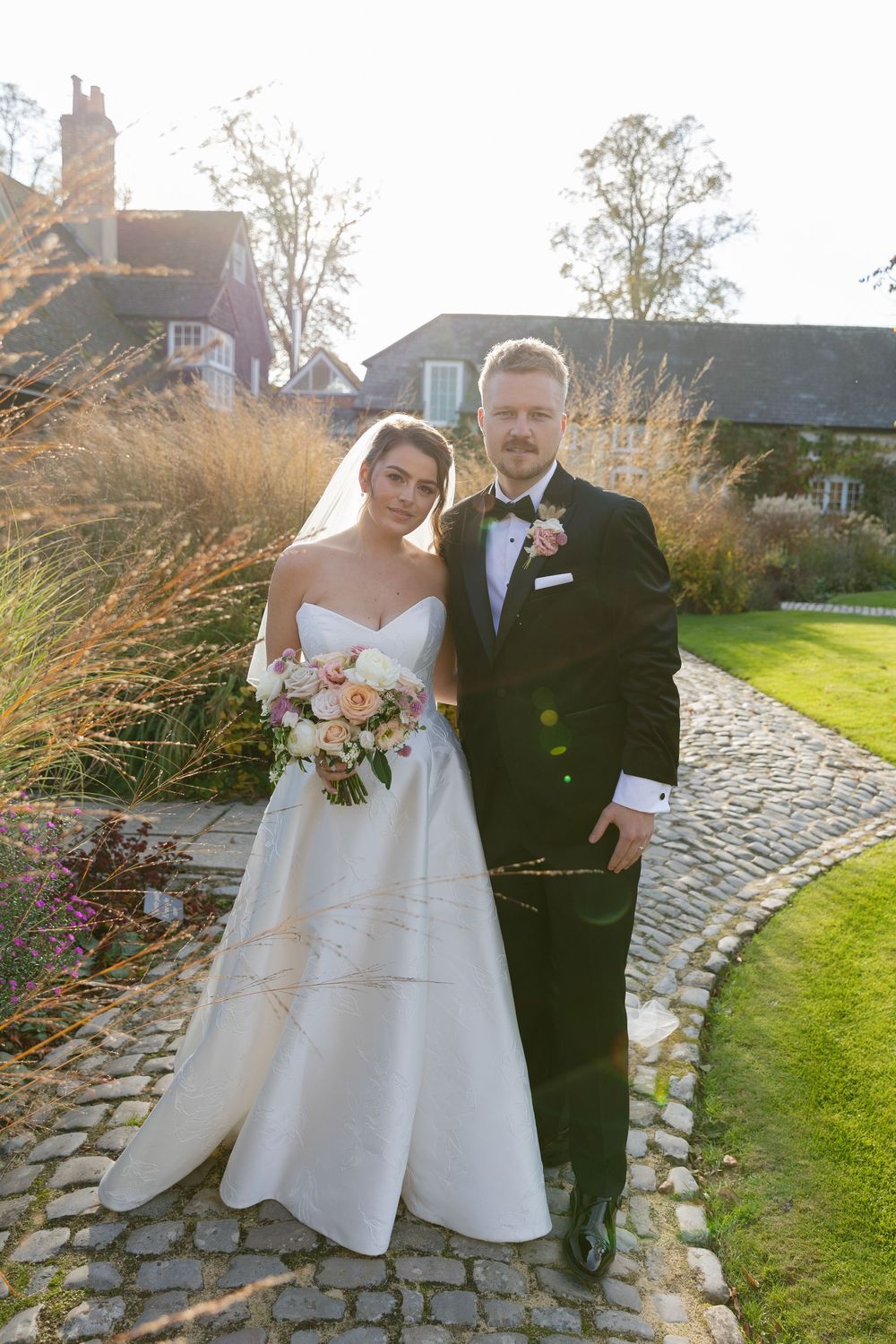 A wedding couple stands on a cobblestone path with ornate gardens and architecture in the background.