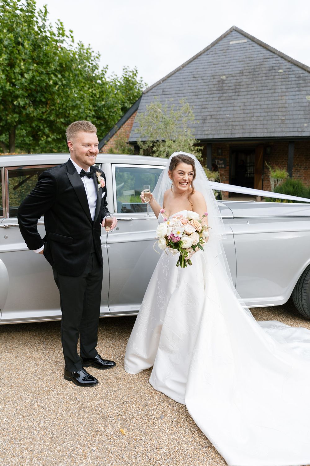 A wedding couple poses beside their classic silver car outside a rustic venue.
