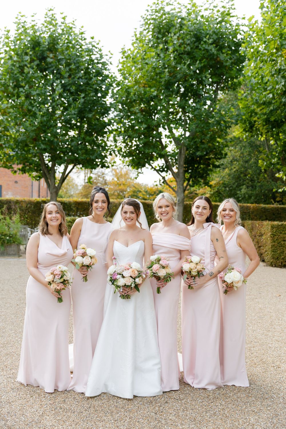 Bridesmaids in soft pink dresses pose with bouquets outdoors during a wedding celebration.