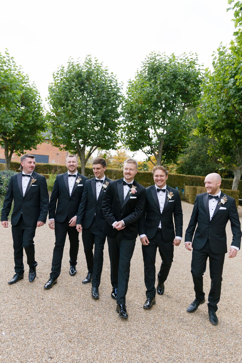 Groomsmen in black tuxedos walking together on a gravel path during wedding celebrations.