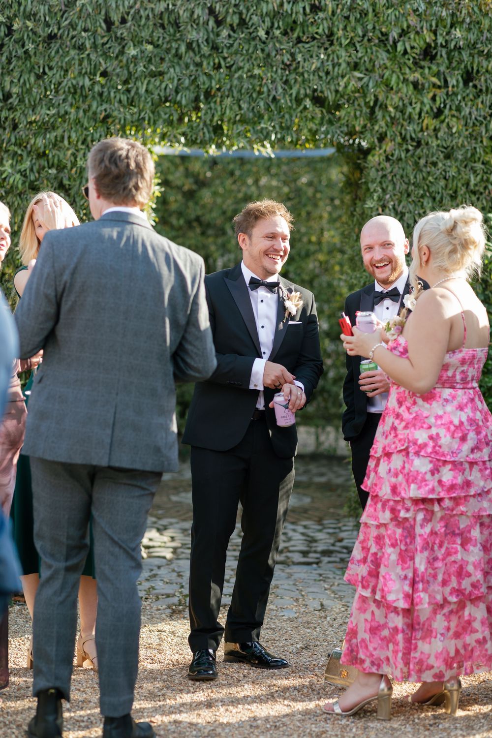 Wedding party socializes on garden path with lush green hedges providing natural backdrop.
