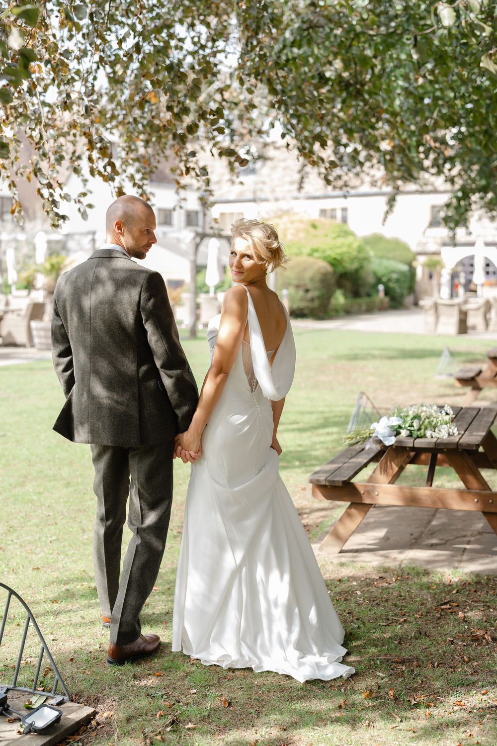 Bride in white gown and groom in gray suit hold hands in a sunlit garden near a wooden picnic table.