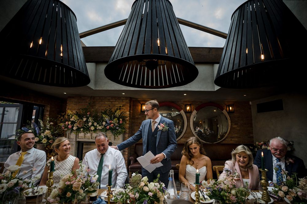 Wedding speeches in modern reception venue with warm ambient lighting and guests laughing