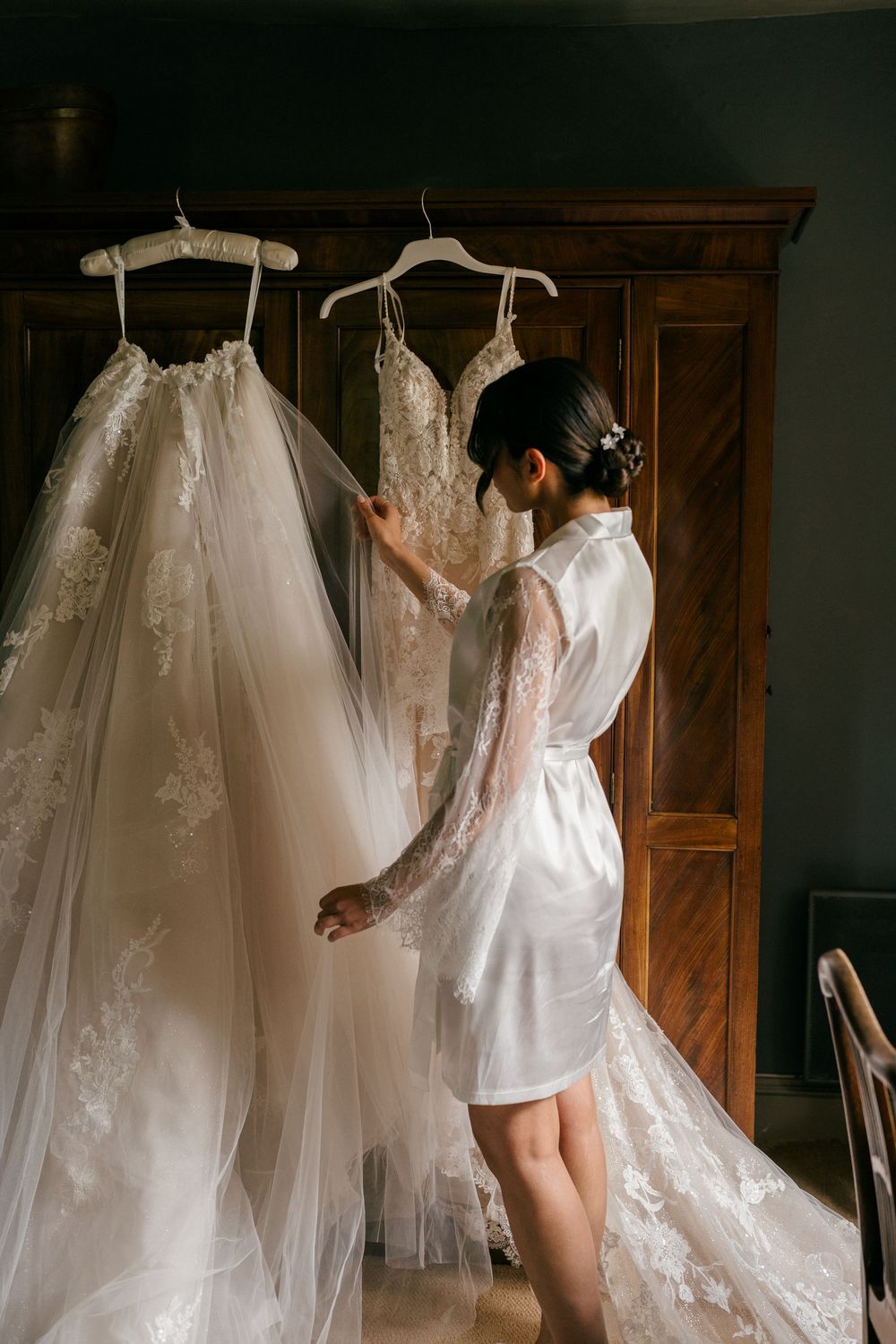 Bride in lace robe admiring wedding dresses hanging on wooden wardrobe in elegant bridal preparation room.