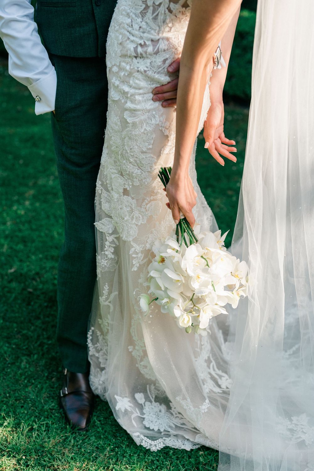 Bride in lace wedding gown holding white floral bouquet beside groom in dark suit on green grass.