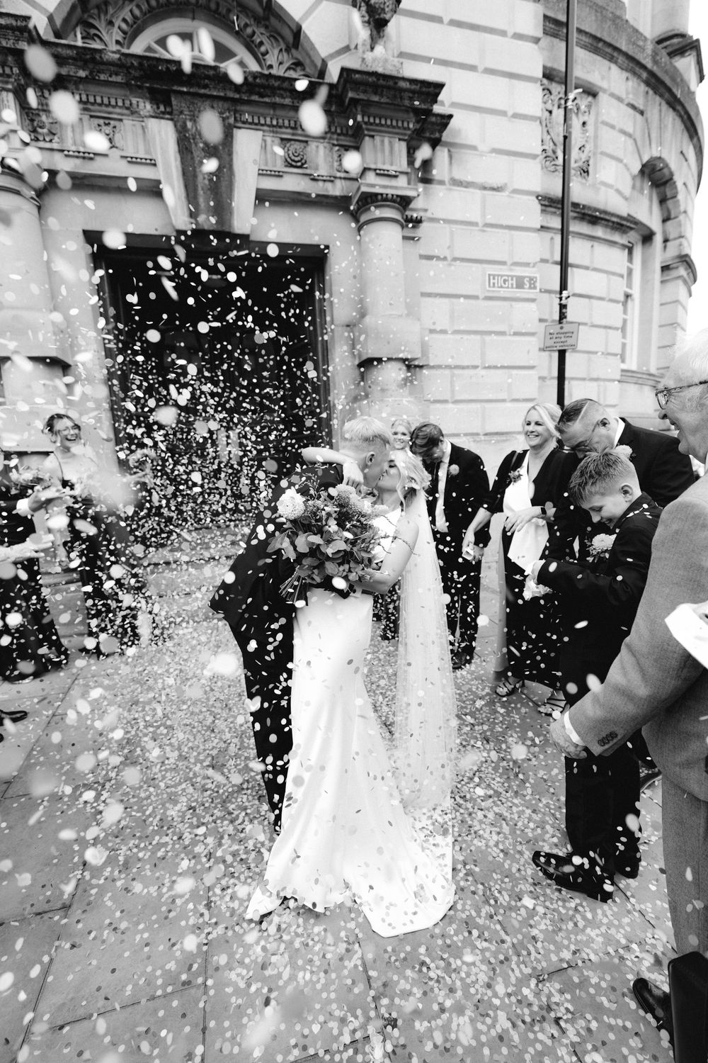 Black and white photo of bride and groom kissing outside church as guests throw confetti.