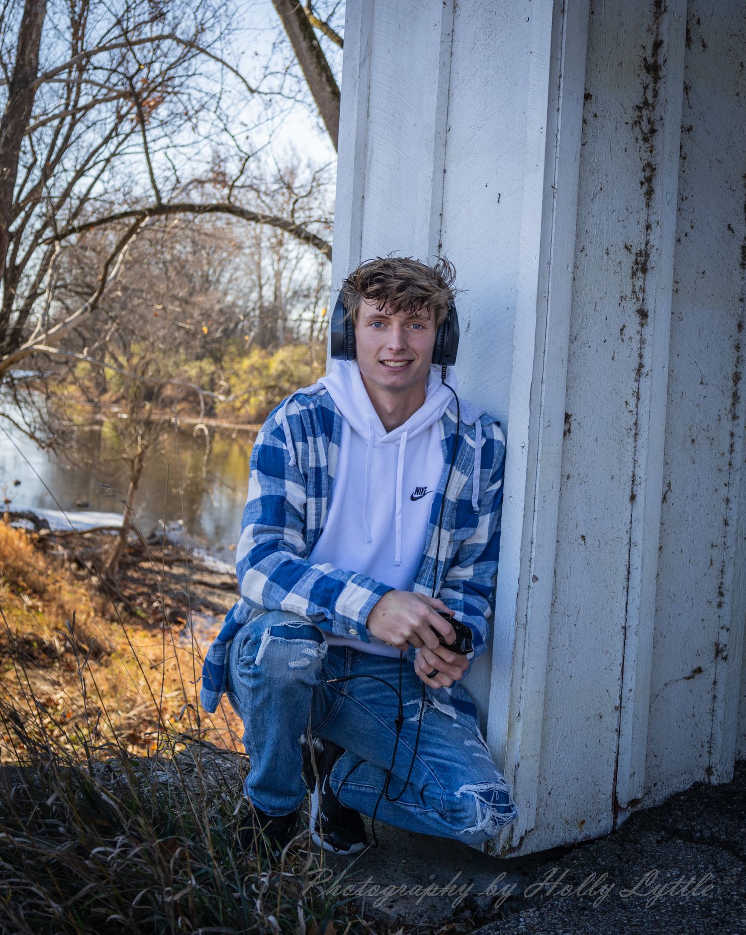 A person in a blue plaid shirt and jeans poses outdoors near a white wall with a scenic pond and bare trees in background.