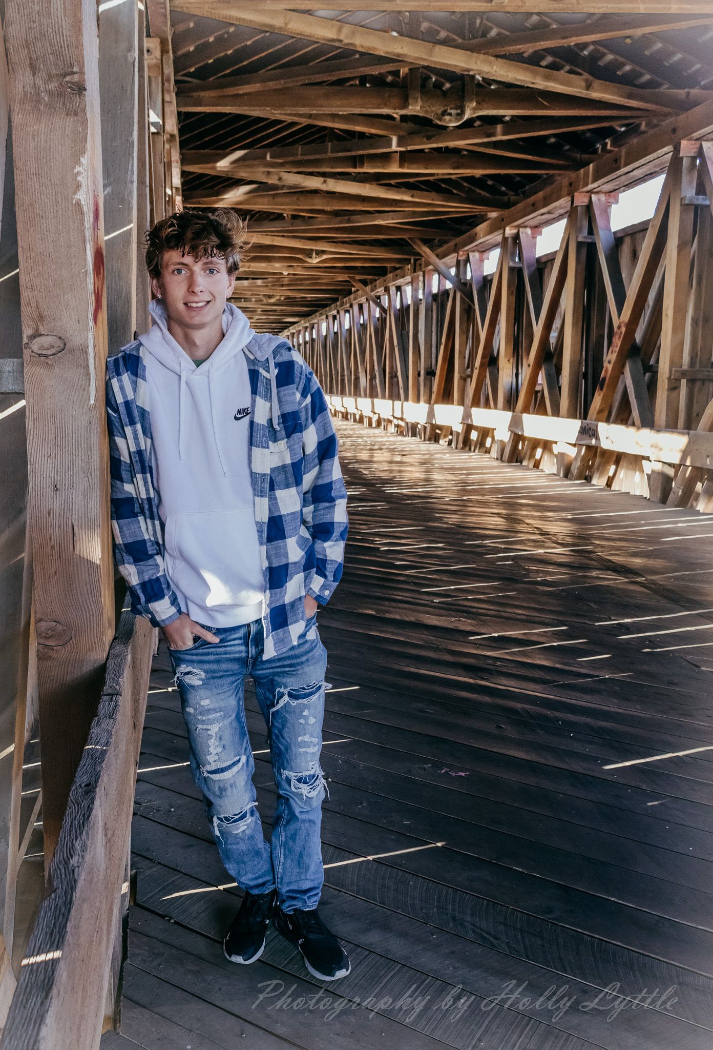 Young person in blue plaid shirt and ripped jeans poses casually against wooden beams in rustic covered bridge interior.
