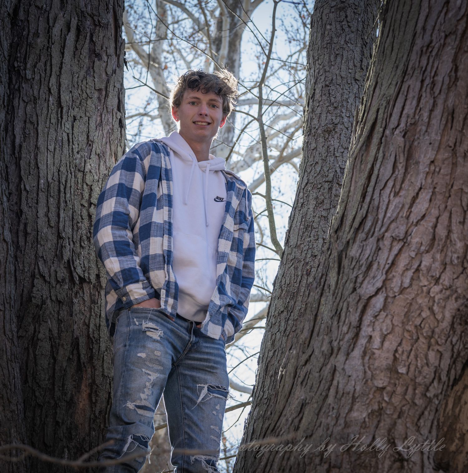 A person in a blue plaid shirt and jeans stands casually by a large tree trunk on a fall day.