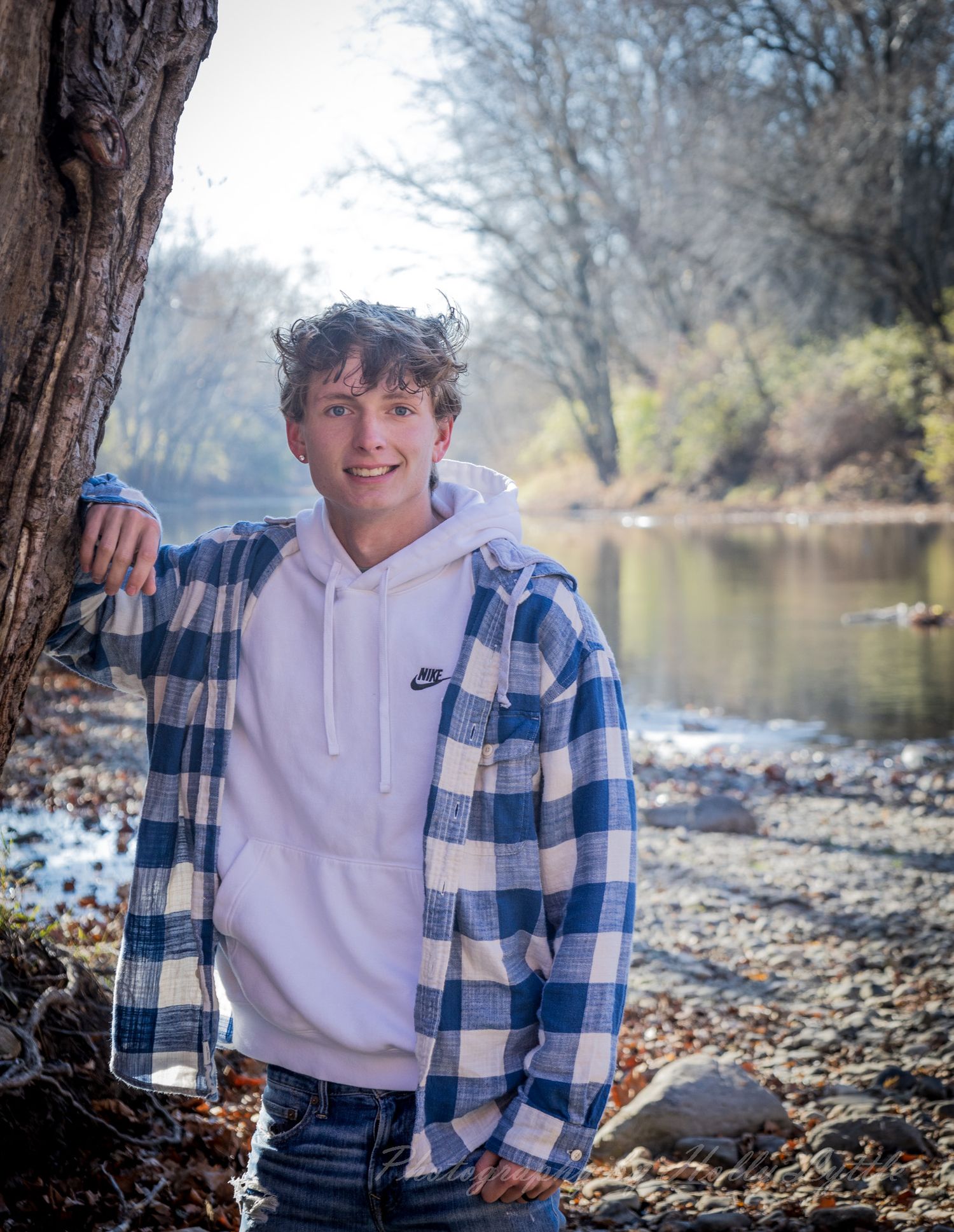 A casual outdoor photoshoot by a river featuring someone wearing a blue plaid flannel and white hoodie.