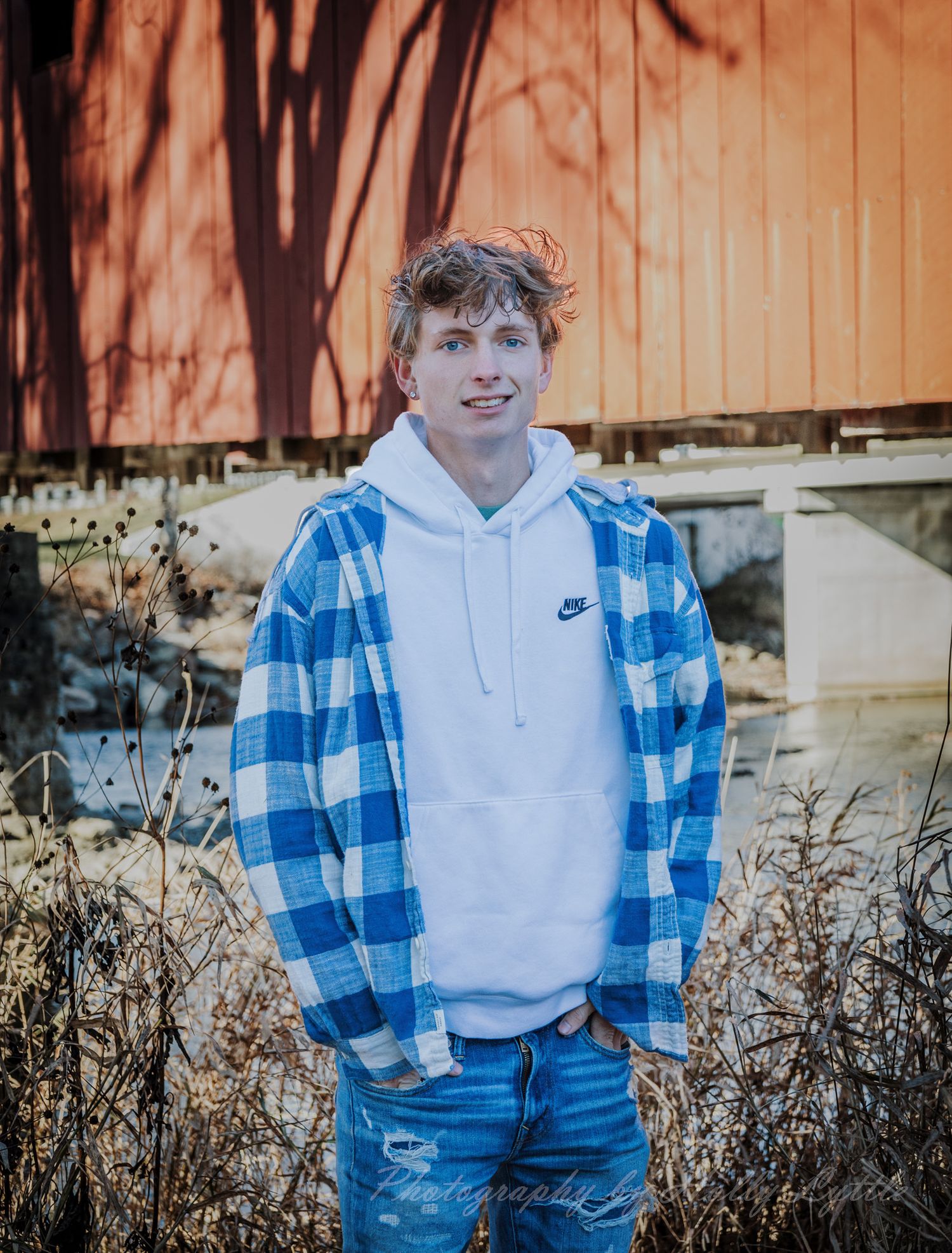 A casual portrait against a wooden wall wearing a blue plaid shirt and white hoodie.