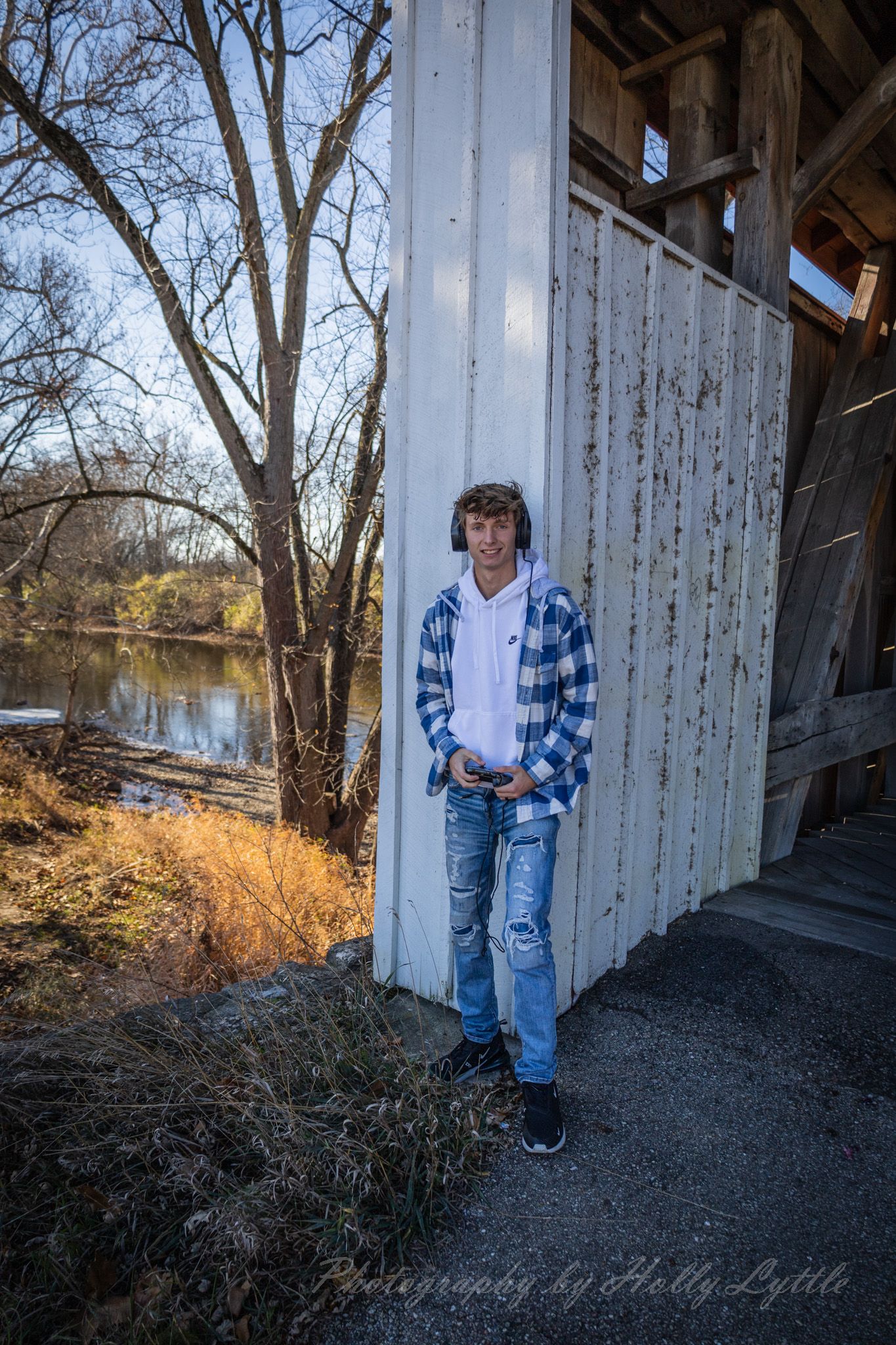 Person in blue plaid flannel and ripped jeans poses against white wall near river and bare trees in natural setting.