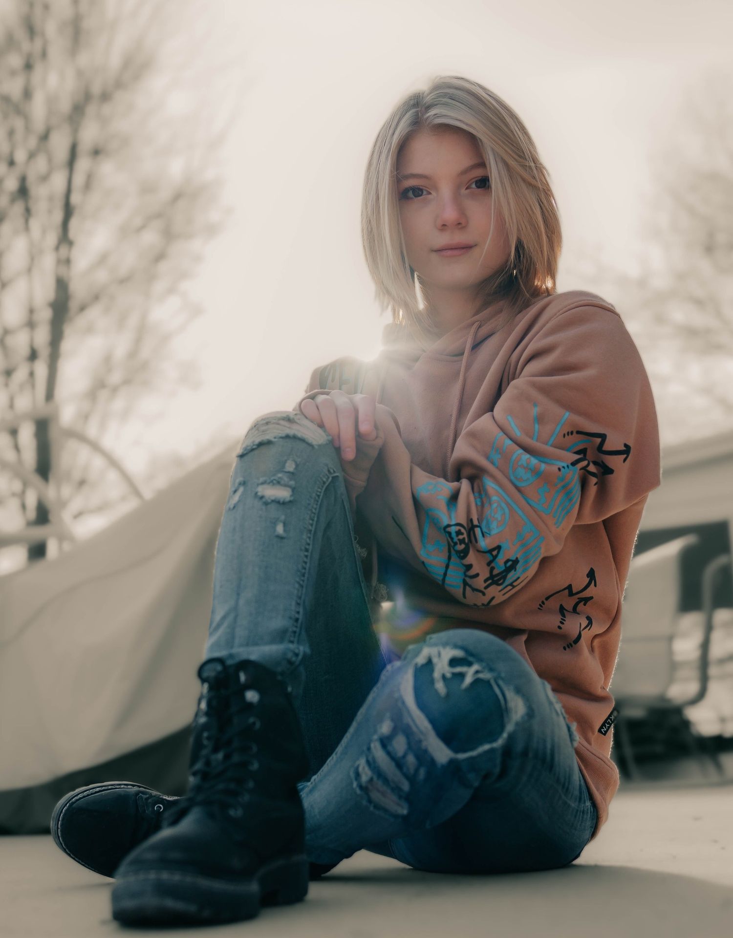 A person in casual attire sits on a snowy ground wearing a brown sweater and jeans in a winter setting.