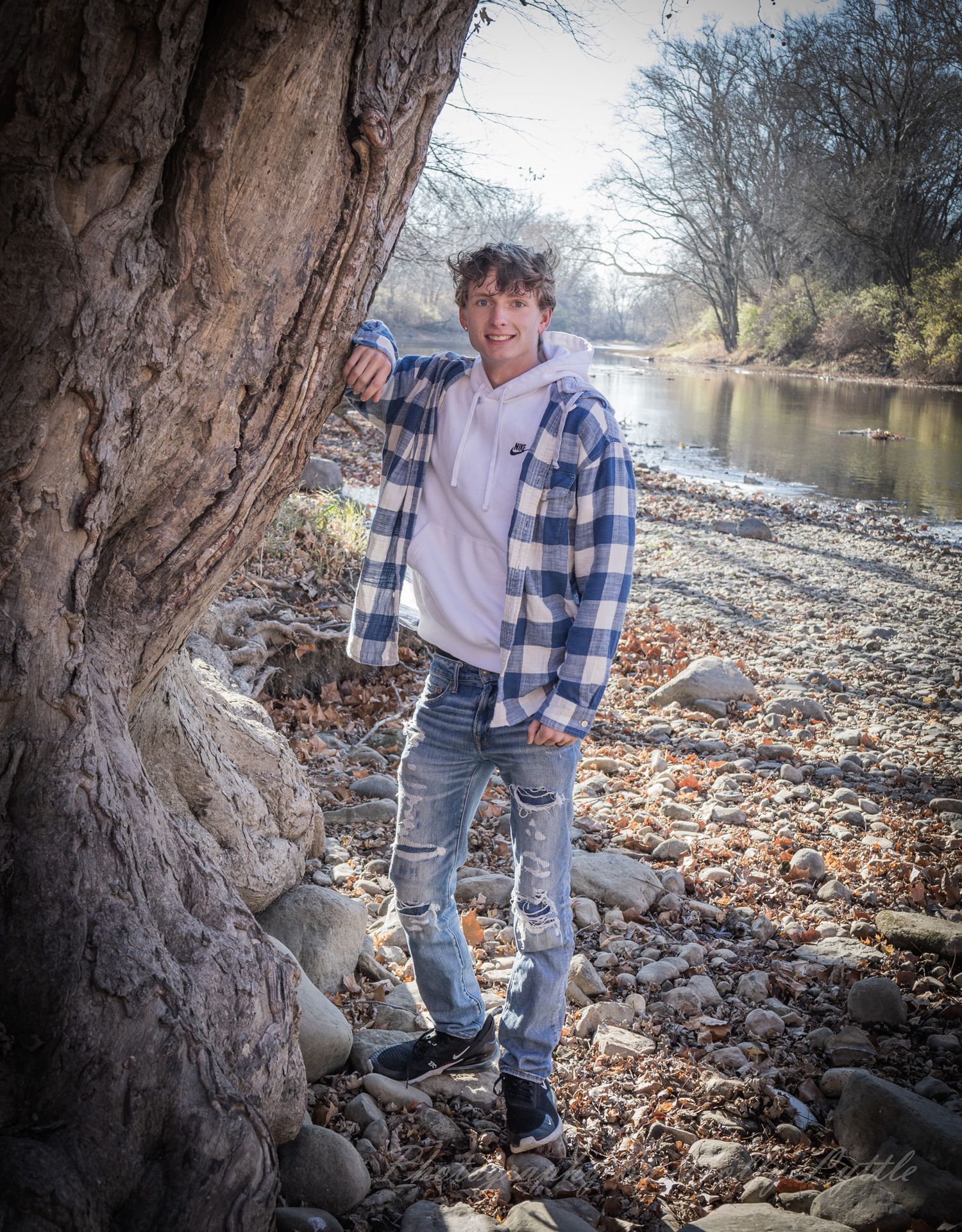 A person in casual attire explores the rocky riverbank surrounded by fallen autumn leaves.