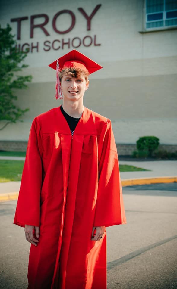 Graduate in bright red cap and gown poses for photos in front of Troy High School on graduation day.