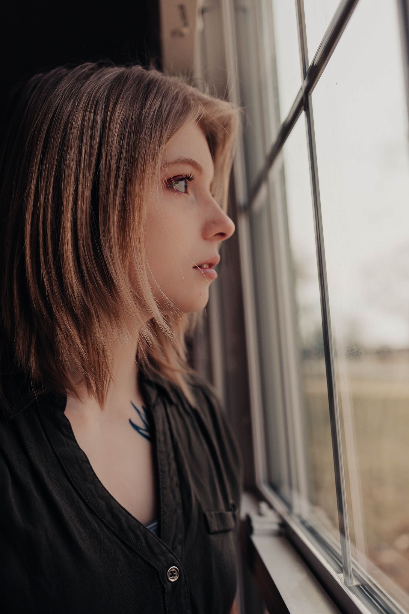 Person with short blonde hair gazing thoughtfully out a window in dramatic lighting.