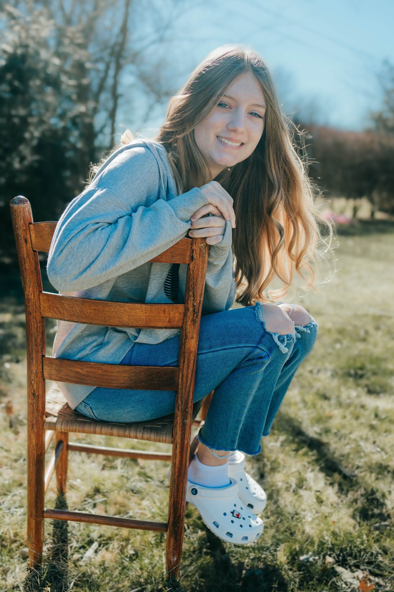 Person in blue sweater and jeans sitting casually on wooden chair outdoors on sunny day.