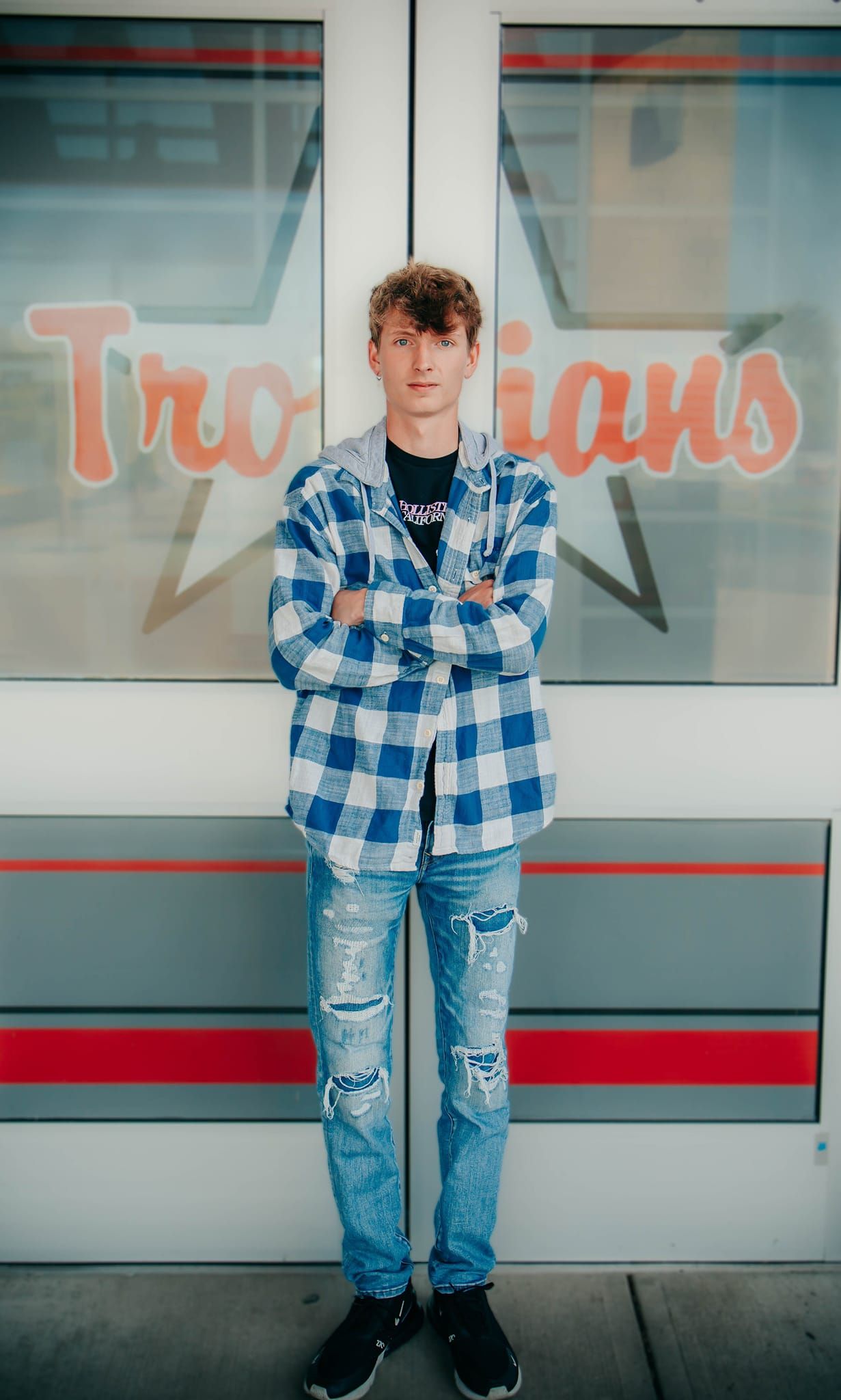 A person in a blue plaid shirt and ripped jeans stands against a storefront window.