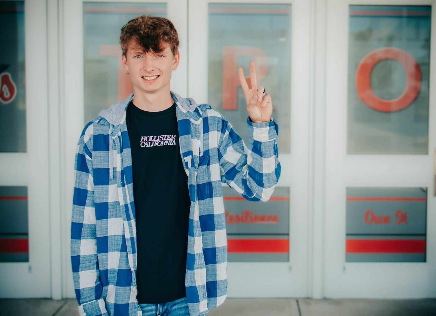 Person in blue plaid shirt makes peace sign in front of storefront windows.